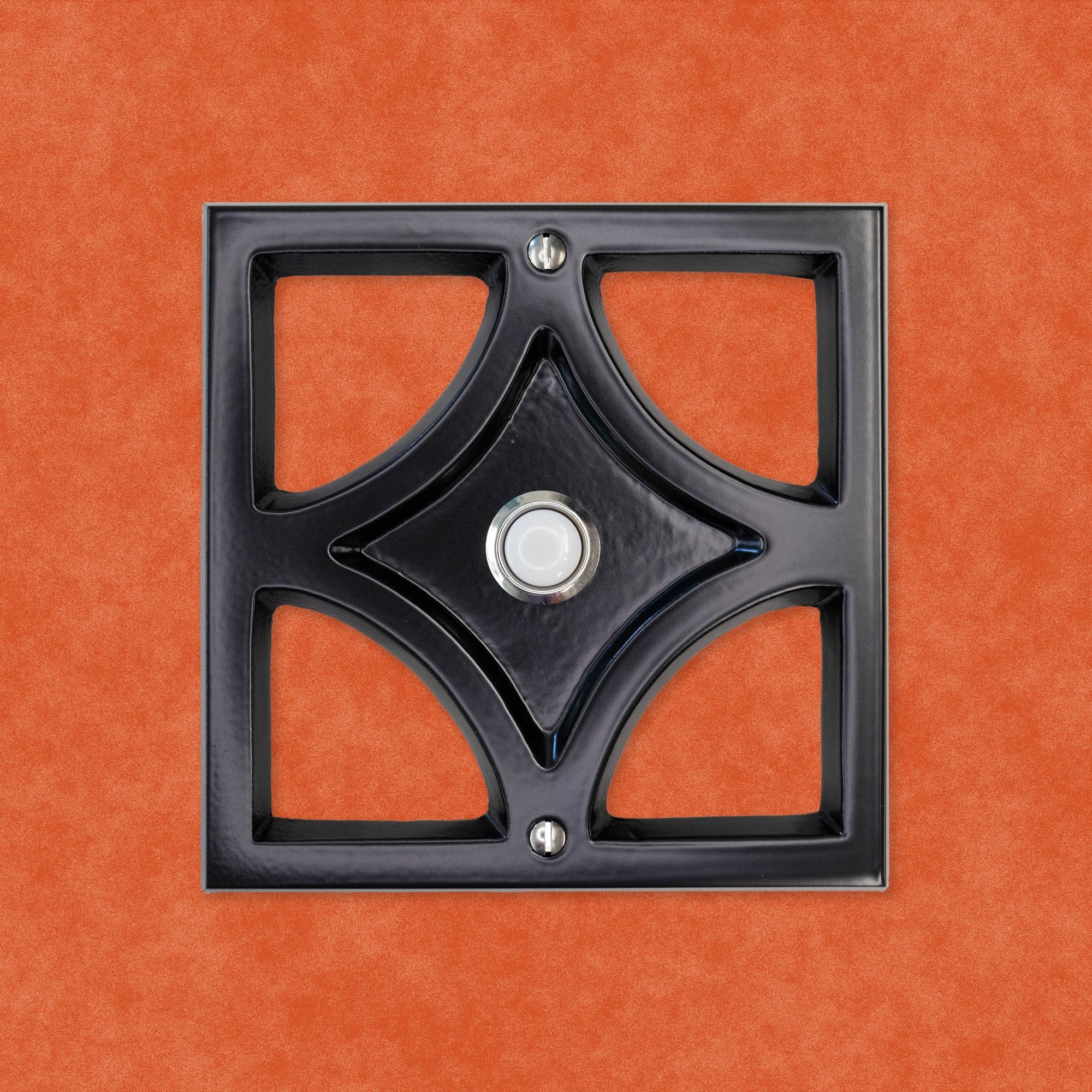 A black powder coated cast aluminum breeze block with a silver rimmed door bell button and screws. The breeze block design is a square around the outside, then inside is a diamond shape leaving 4 hollow corners. The middle of the diamond is sunken in and that is where the doorbell button is.  There are two silver screws, one at the top and bottom of the square. The black powder coating is matte, so it is smooth and reflects light but is does not give a glossy shine.