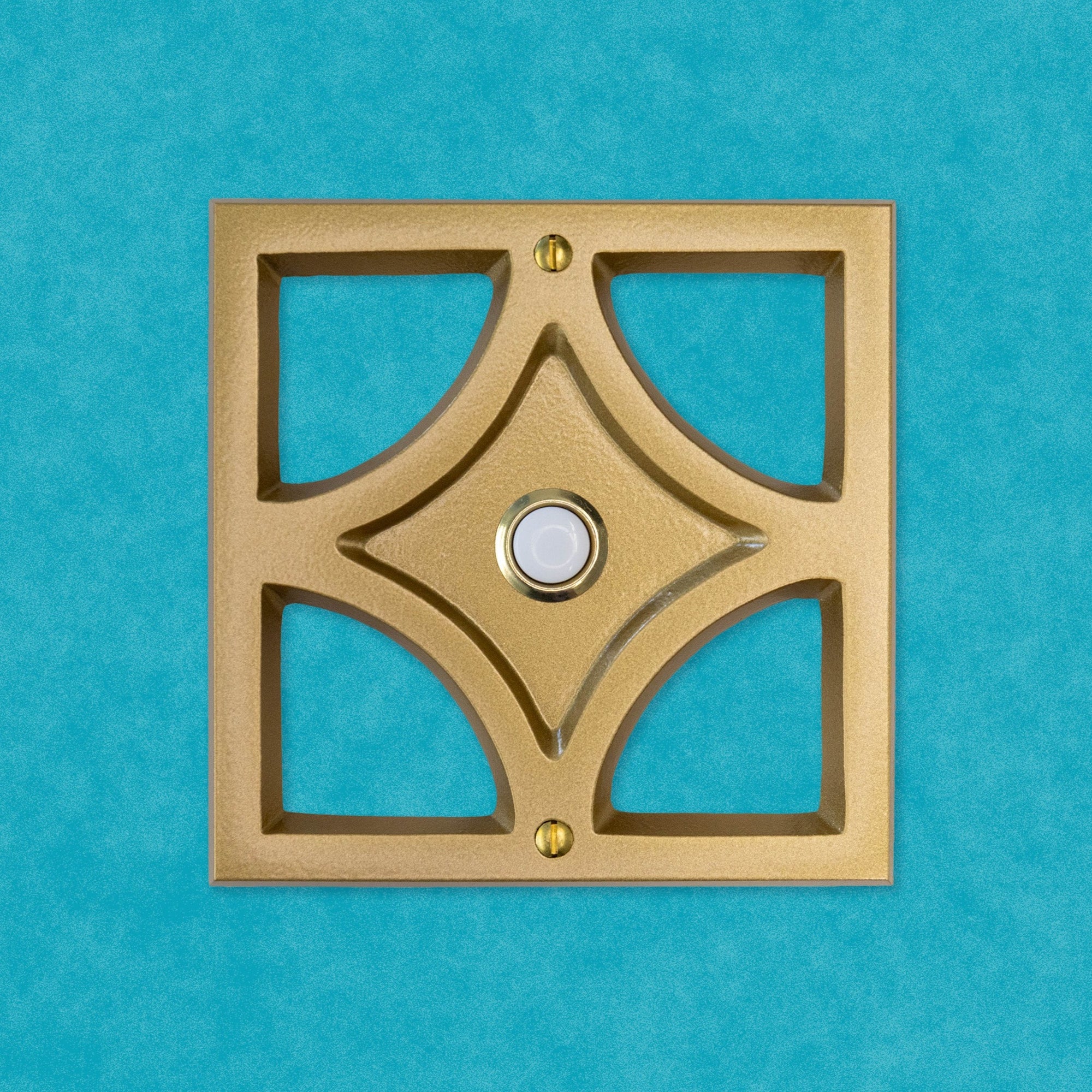 A gold powder coated cast aluminum breeze block with a gold rimmed door bell button and screws. The breeze block design is a square exterior with a diamond shape inside, leaving 4 hollow corners. The middle of the diamond is sunken in, and that is where the doorbell button is. There are two gold screws, one at the top and bottom of the square. The gold powder coating is smooth in texture and has a bit of reflective shine to it