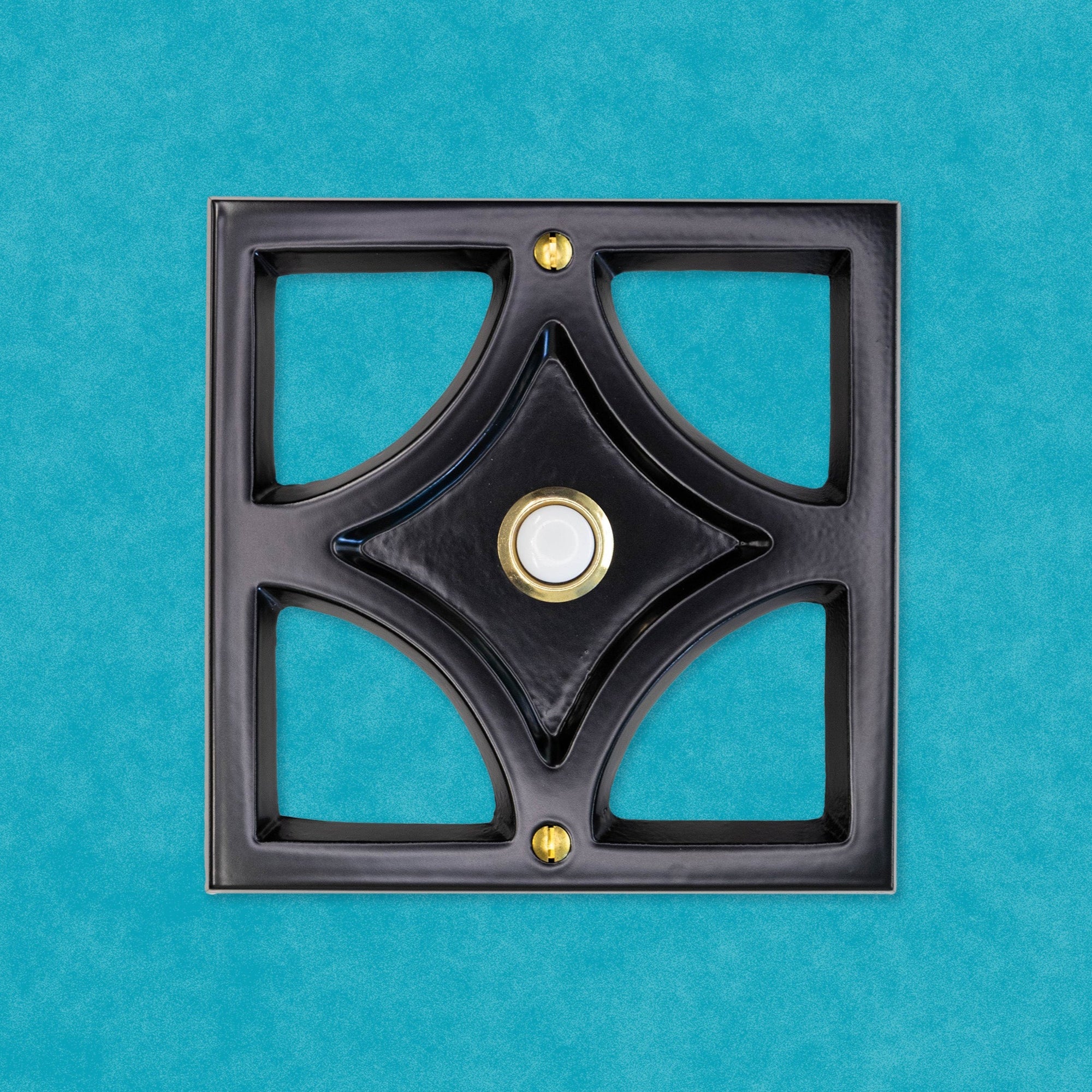 A black powder coated cast aluminum breeze block with a gold rimmed door bell button and screws. The breeze block design is a square around the outside, then inside is a diamond shape leaving 4 hollow corners. The middle of the diamond is sunken in and that is where the doorbell button is. There are two gold screws, one at the top and bottom of the square. The black powder coating is matte, so it is smooth and reflects light but is does not give a glossy shine.