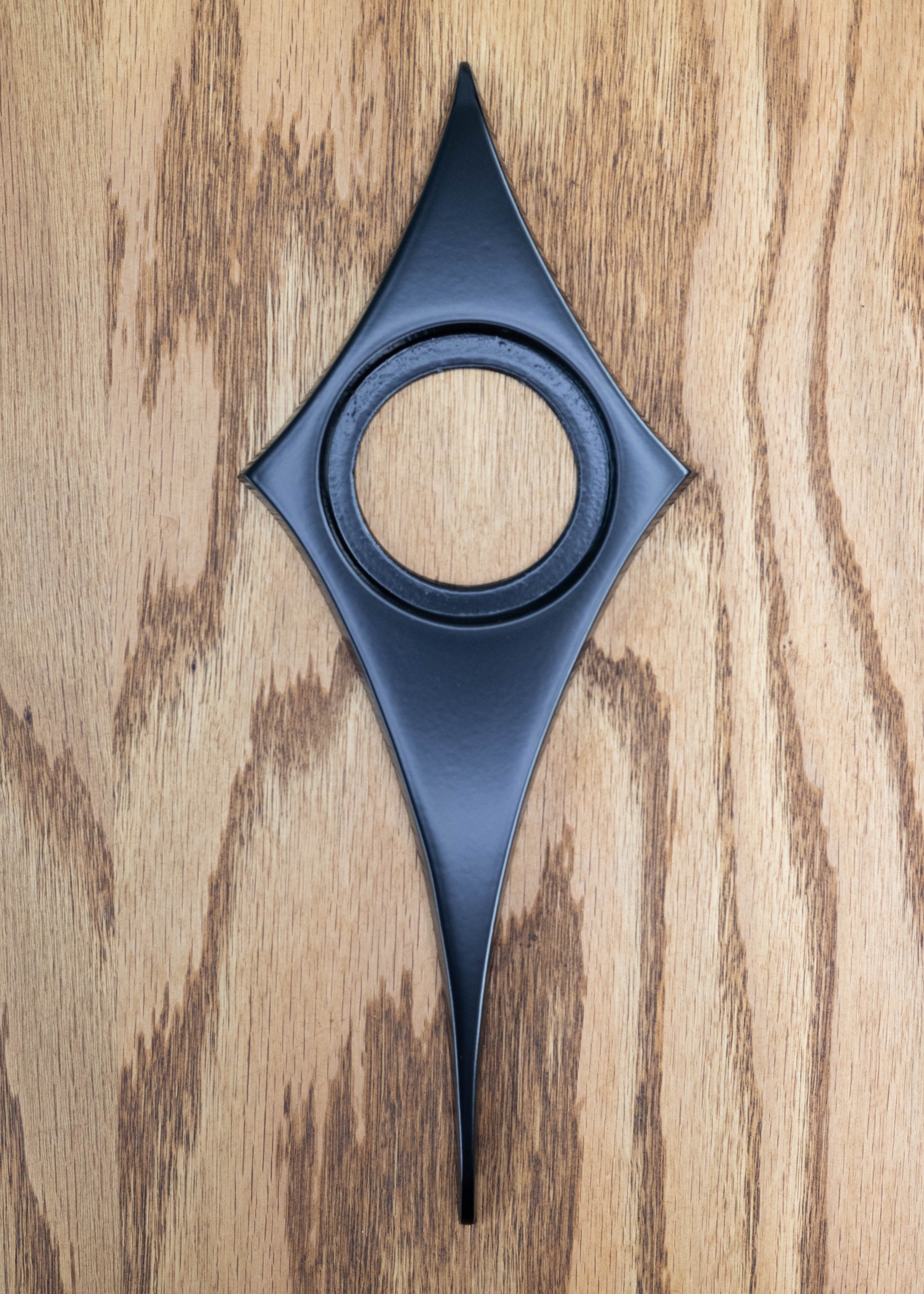 Matte black powder coated comet doorknob escutcheon. The surface is very smooth, and reflects light but is not glossy. The comet shape is like a diamond with slightly curved in edges and the bottom point is long. There is a hole in the center where the door knob goes.