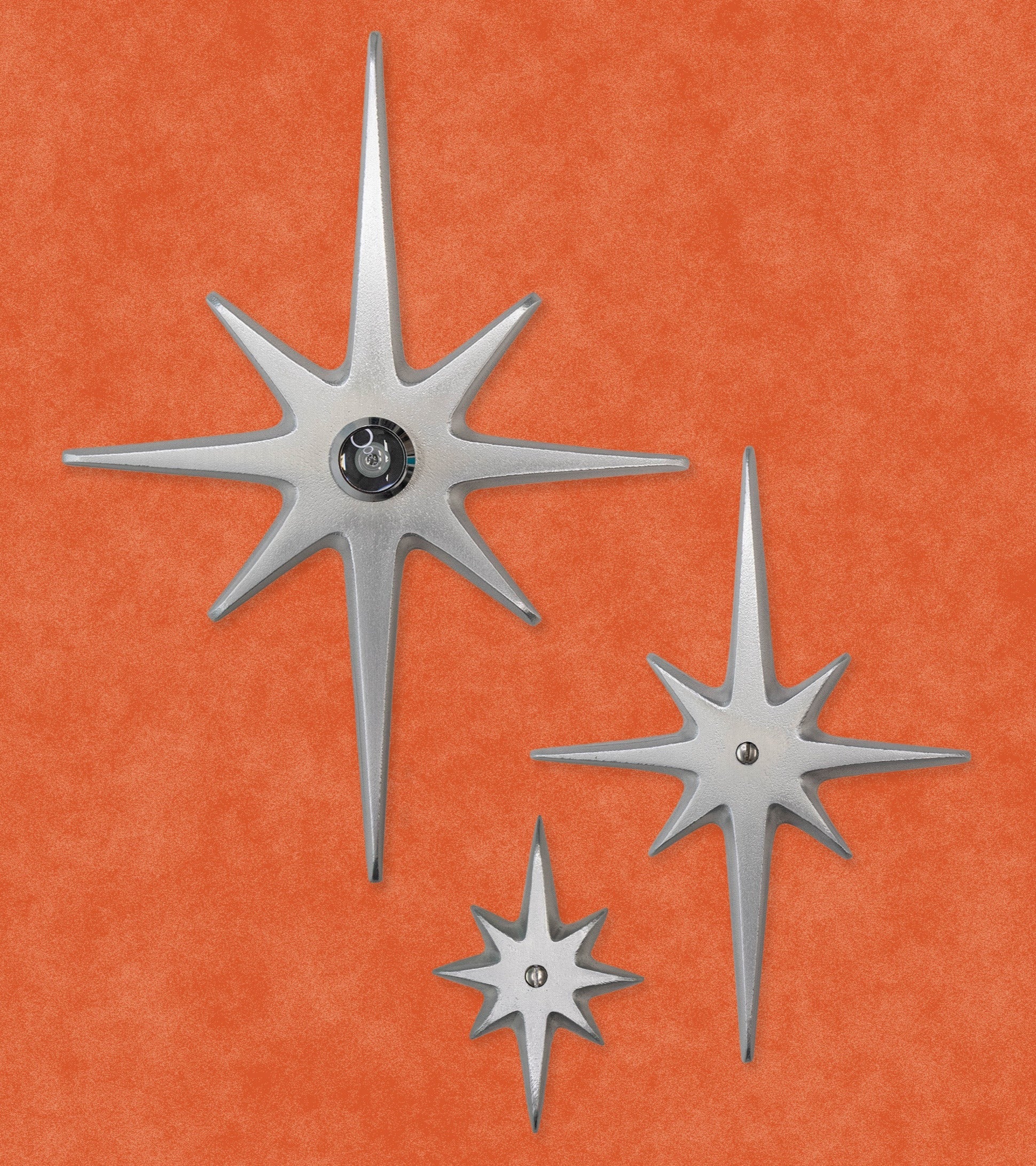A set of three cast aluminum starbursts: Large, medium, and small. The largest one has a peephole viewer in the center, while the other two have a single screw at their centers. The style of starburst has the 2 longest points top and bottom, 2 medium points horizontal, and the other 4 shortest points diagonal in between. The aluminum is silver in color. They have a smooth but slightly textured finish due to the natural aluminum. There is slight shine, but are not glossy.