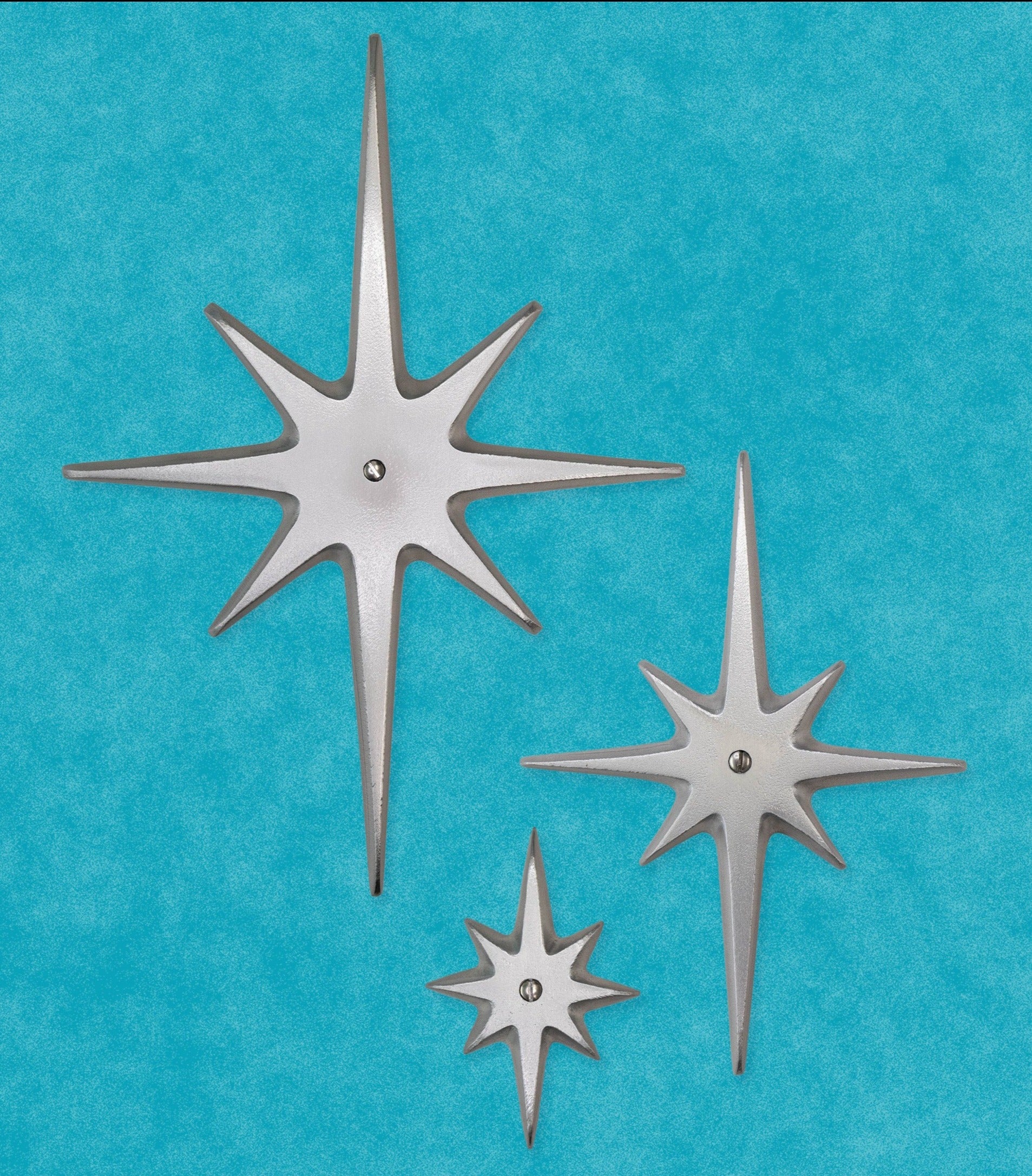 A set of three cast aluminum starbursts: Large, medium, and small, each with a single screw at the very center. The style of starburst has the 2 longest points top and bottom, 2 medium points horizontal, and the other 4 shortest points diagonal in between. The aluminum is silver in color. They have smooth but slightly textured finish due to the natural aluminum. There is slight shine, but are not glossy.