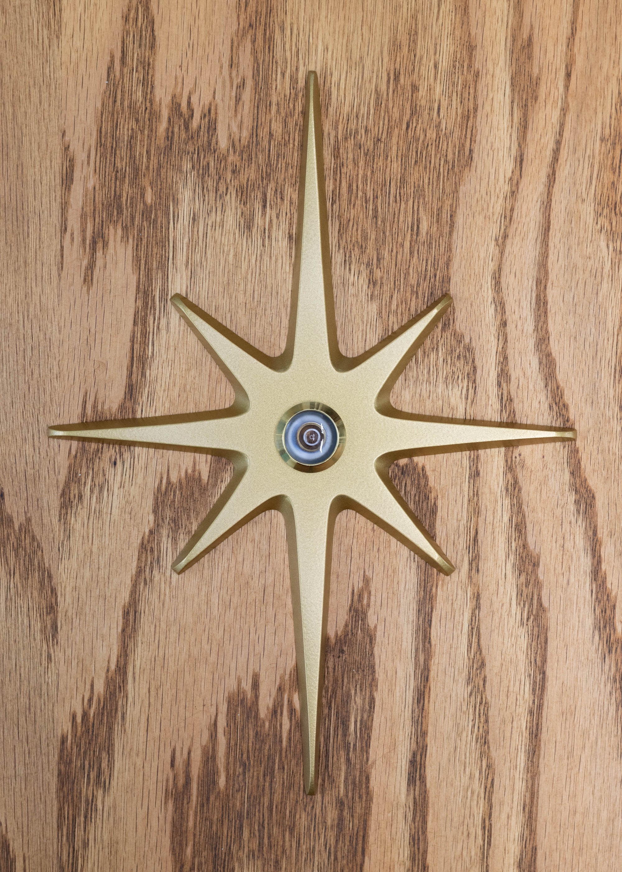 A large cast aluminum starburst with a peephole viewer at its center. It is gold powder coated with a gold rimmed peephole. The powder coating provides a smooth finish with slight metallic shine to it.