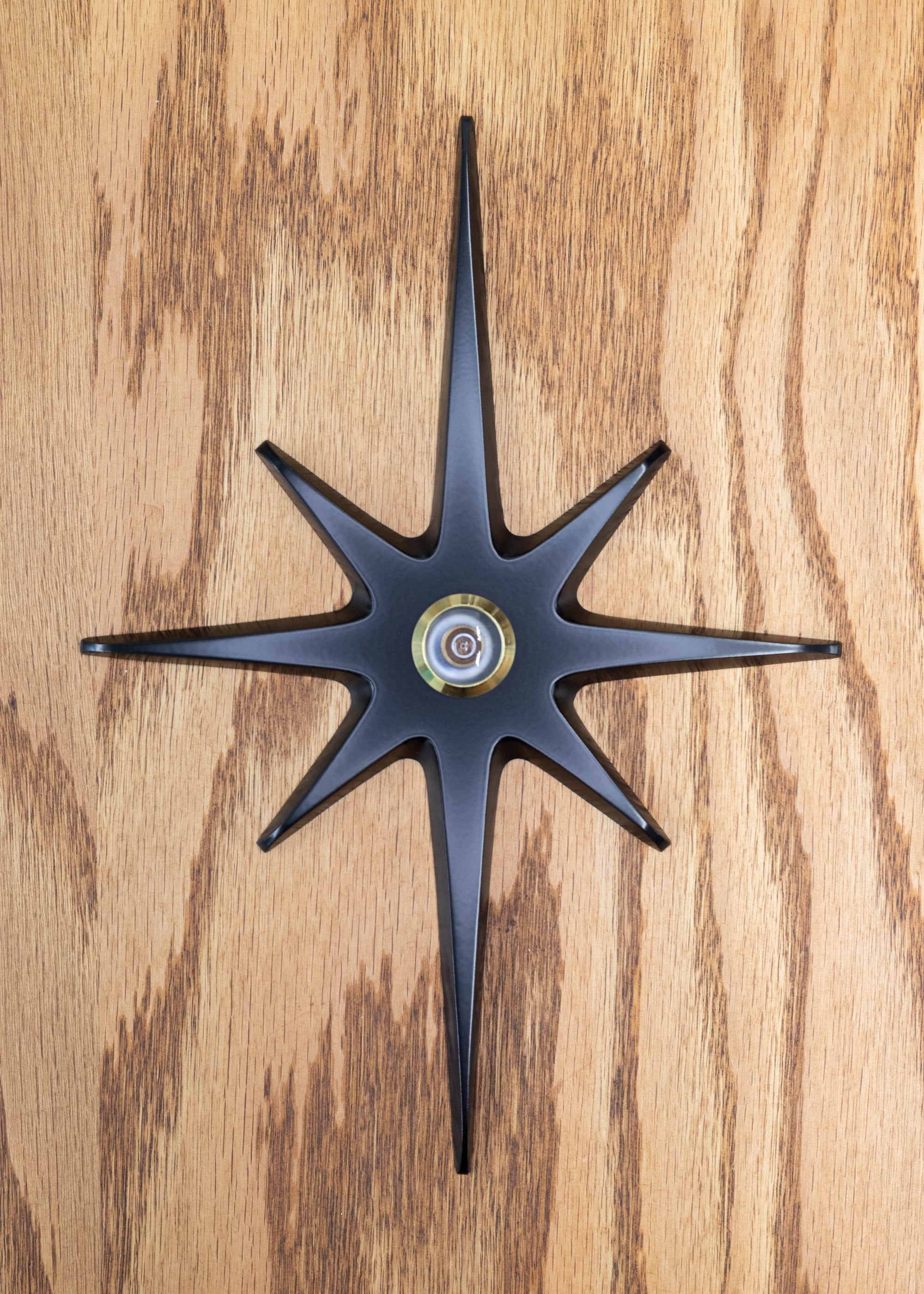 A large cast aluminum starburst with a peephole viewer in the center. The starburst has a black powder coating and the peephole is gold rimmed. The powder coat finish is matte, so it's not glossy with shine, but the surface is still smooth and gives slight reflection of light.