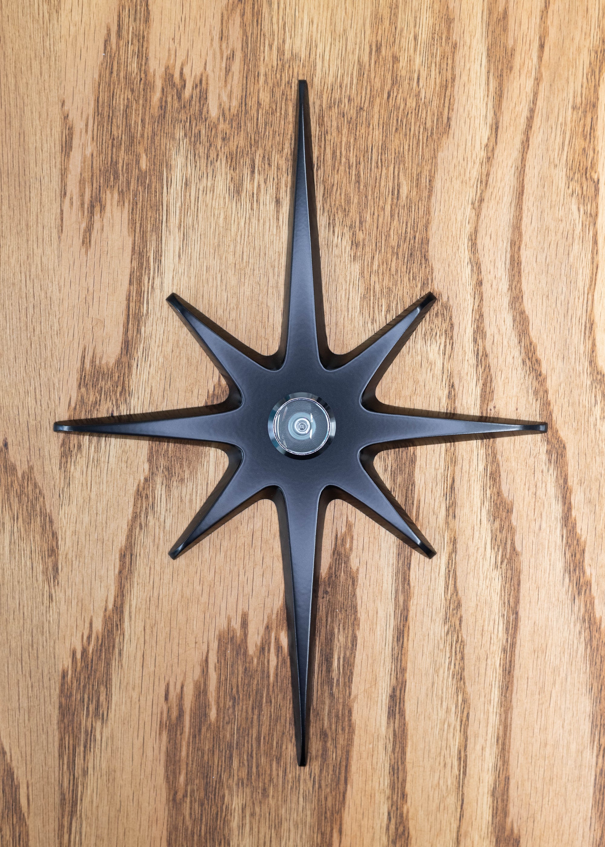 A large cast aluminum starburst with a peephole viewer in the center. The starburst has a black powder coating and the peephole is silver/chrome rimmed. The powder coat finish is matte, so it's not glossy with shine, but the surface is still smooth and gives slight reflection of light.