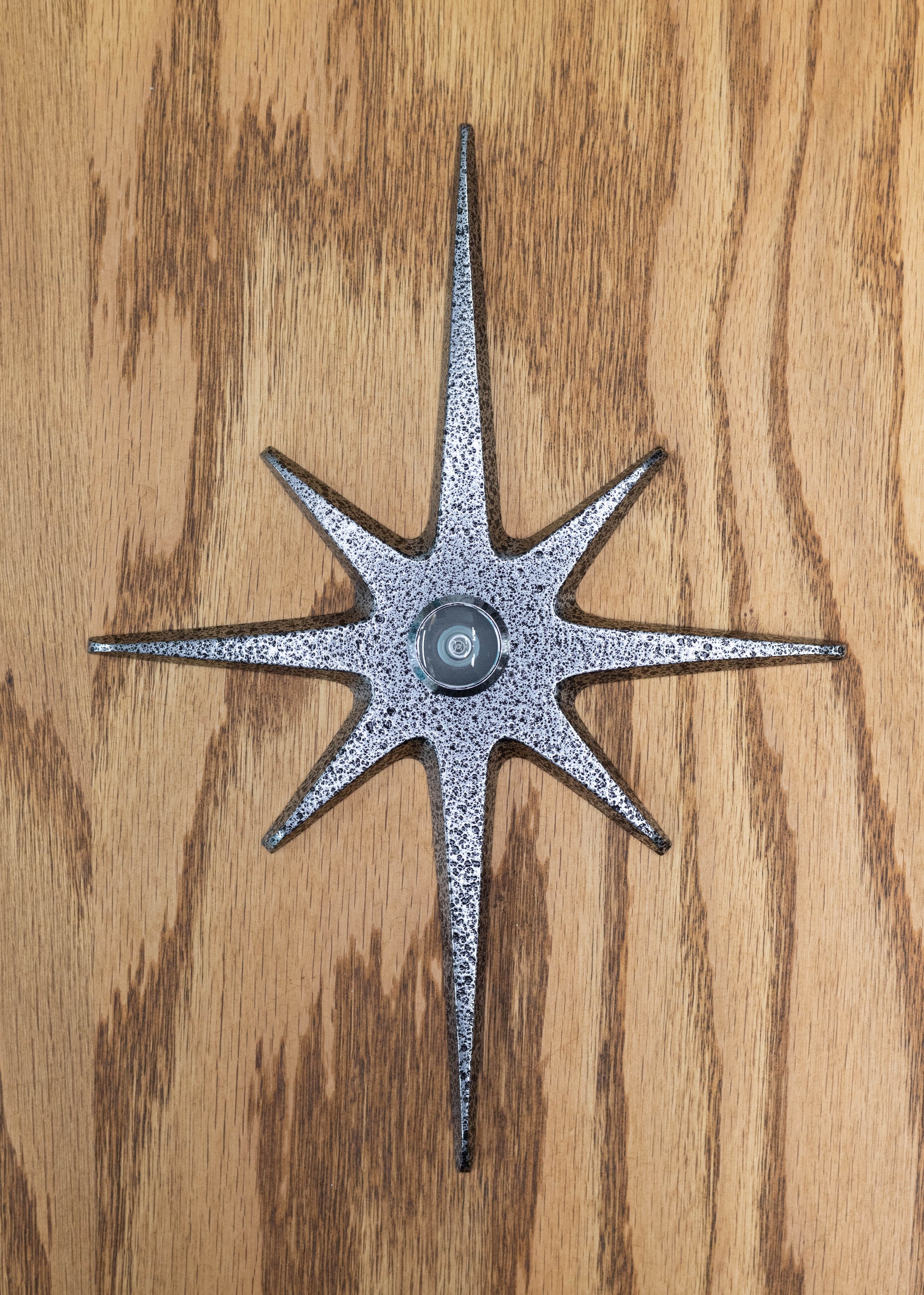 A large cast aluminum starburst with a peephole viewer at the center. The starburst has a black frost powder coating which is a silver and black crackled and pitted texture. The peephole is silver/chrome.