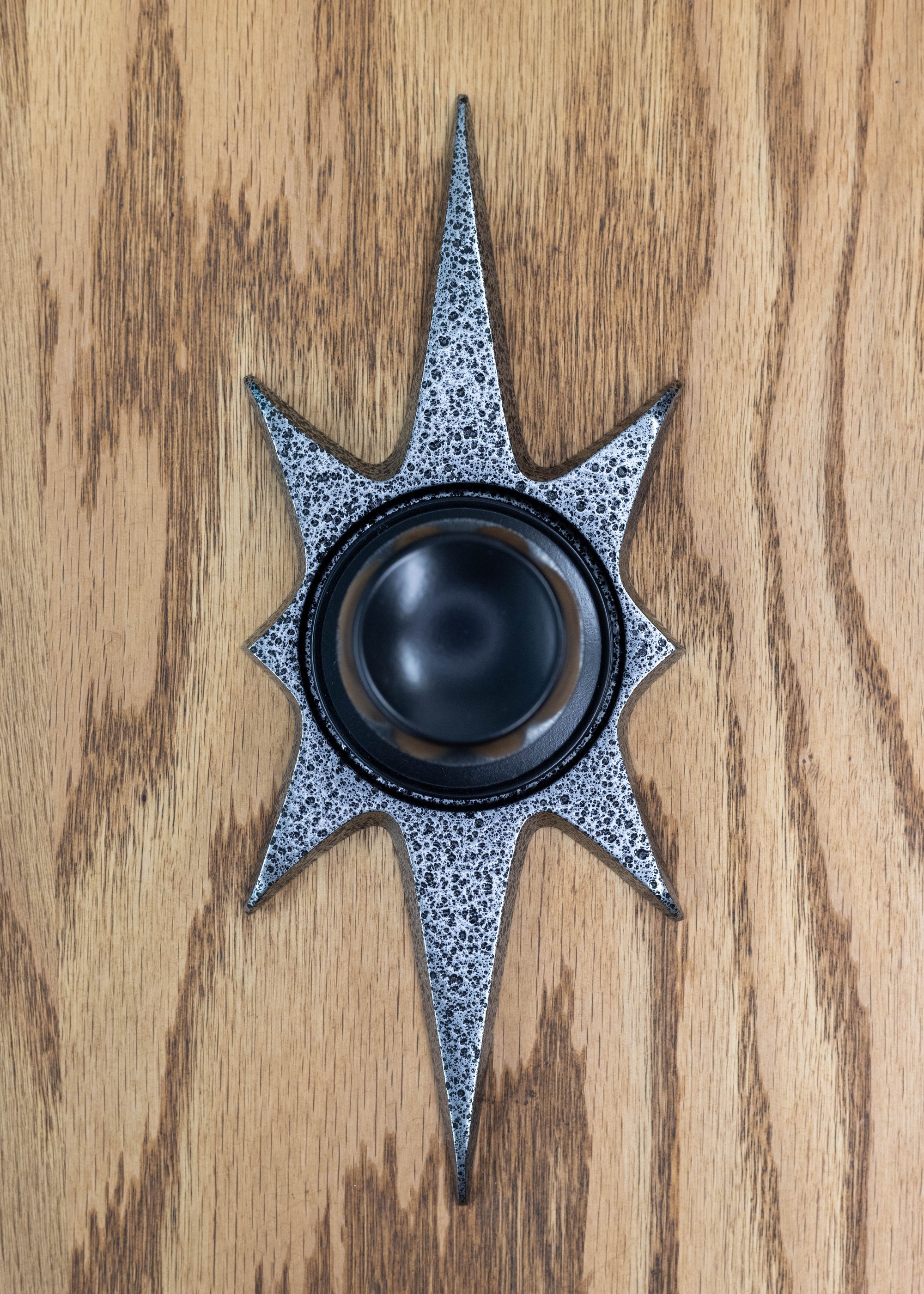 A starburst doorknob escutcheon with a black door knob. (knob not for sale) It is a dark silver, almost gunmetal color with a porous/pitted texture that is black. The starburst consists of two long points on top and bottom, 2 very short points horizontal, and 4 medium points diagonal.