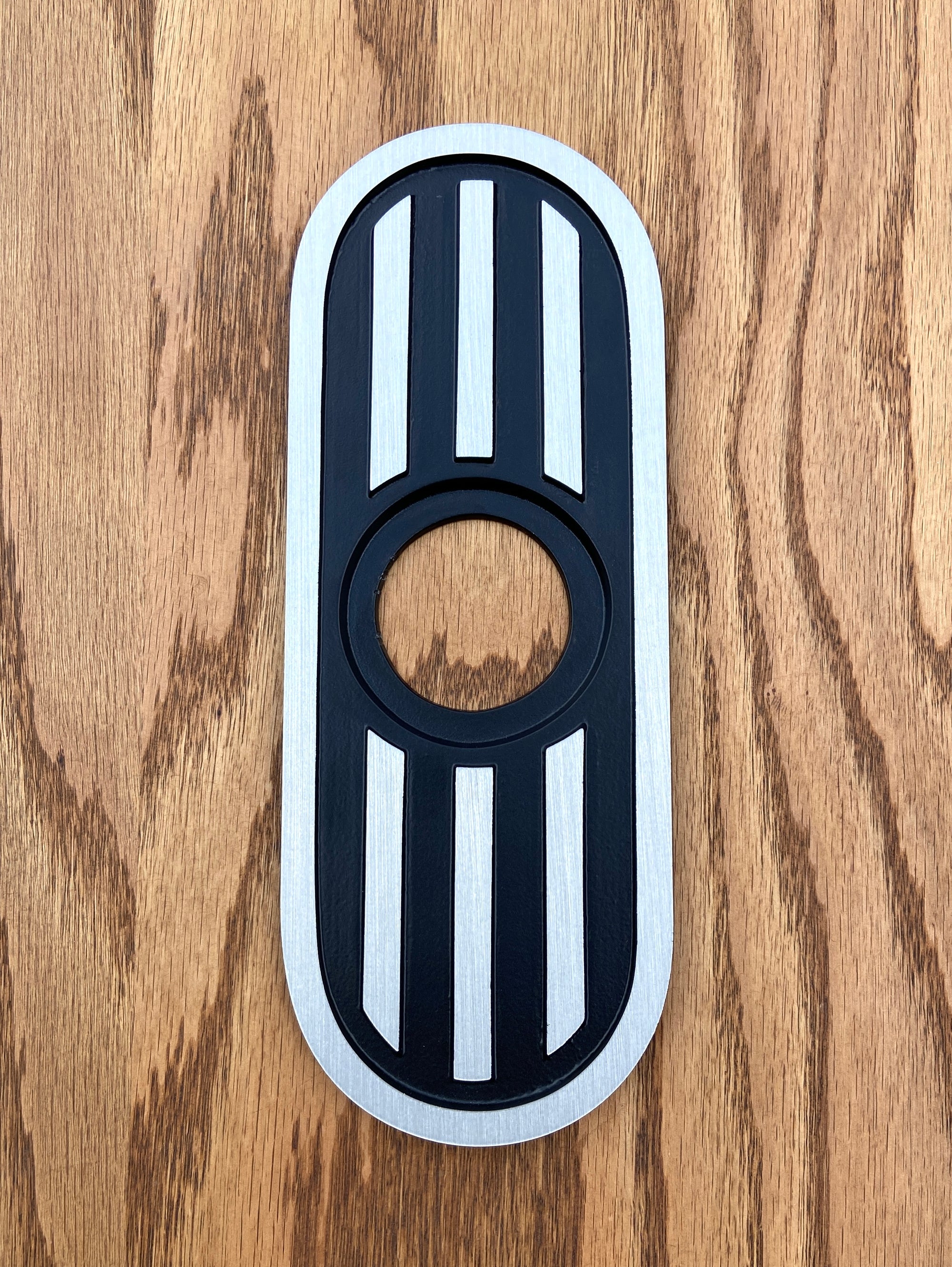 A cast aluminum door knob escutcheon. It is a long vertical oval shape, with three vertical stripes in the middle. There is a hole in the very center where the doorknob goes. The outer edge  and the three vertical stripes are raised from the middle sunken area to give contrast.