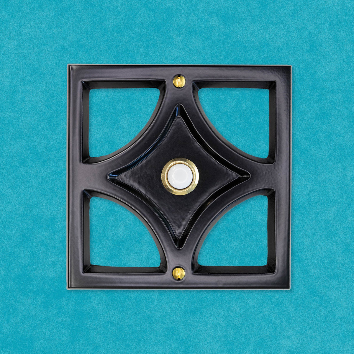 A black powder coated cast aluminum breeze block with a gold rimmed door bell button and screws. The breeze block design is a square around the outside, then inside is a diamond shape leaving 4 hollow corners. The middle of the diamond is sunken in and that is where the doorbell button is. There are two gold screws, one at the top and bottom of the square. The black powder coating is matte, so it is smooth and reflects light but is does not give a glossy shine.