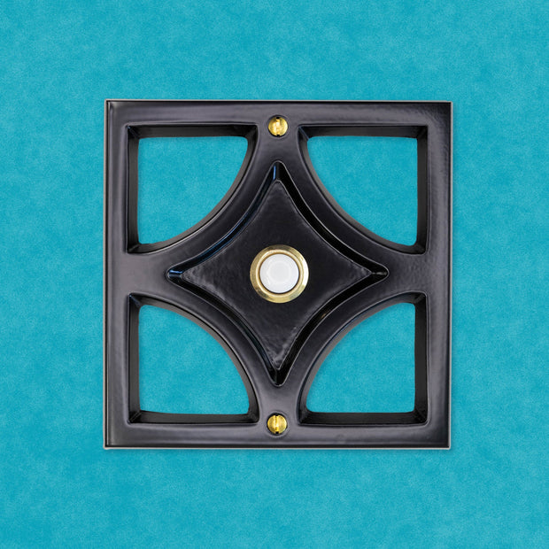 A black powder coated cast aluminum breeze block with a gold rimmed door bell button and screws. The breeze block design is a square around the outside, then inside is a diamond shape leaving 4 hollow corners. The middle of the diamond is sunken in and that is where the doorbell button is. There are two gold screws, one at the top and bottom of the square. The black powder coating is matte, so it is smooth and reflects light but is does not give a glossy shine.