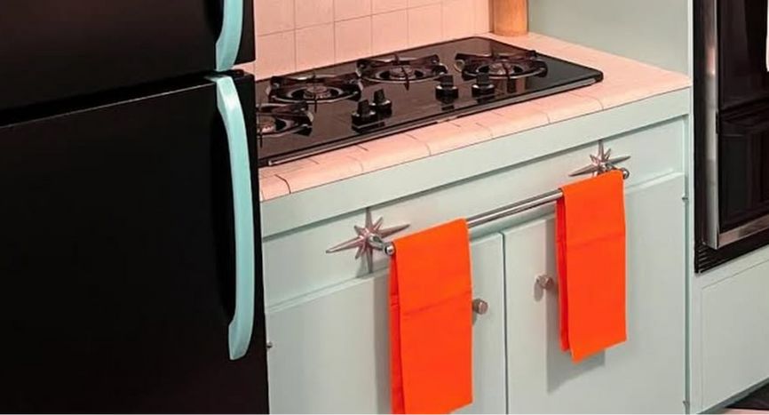 A retro-style kitchen scene featuring a black refrigerator with light blue handles, a black gas stovetop, a pale mint green cabinet with orange towels hanging from Atomic Starburst towel bar, and a pink tiled backsplash.