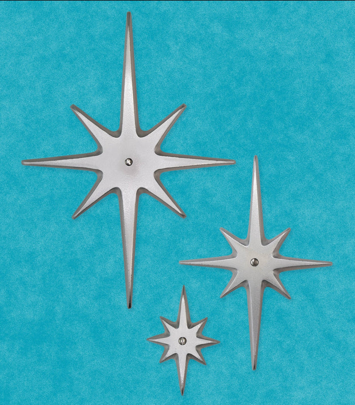 A set of three cast aluminum starbursts: Large, medium, and small, each with a single screw at the very center. The style of starburst has the 2 longest points top and bottom, 2 medium points horizontal, and the other 4 shortest points diagonal in between. The aluminum is silver in color. They have smooth but slightly textured finish due to the natural aluminum. There is slight shine, but are not glossy.