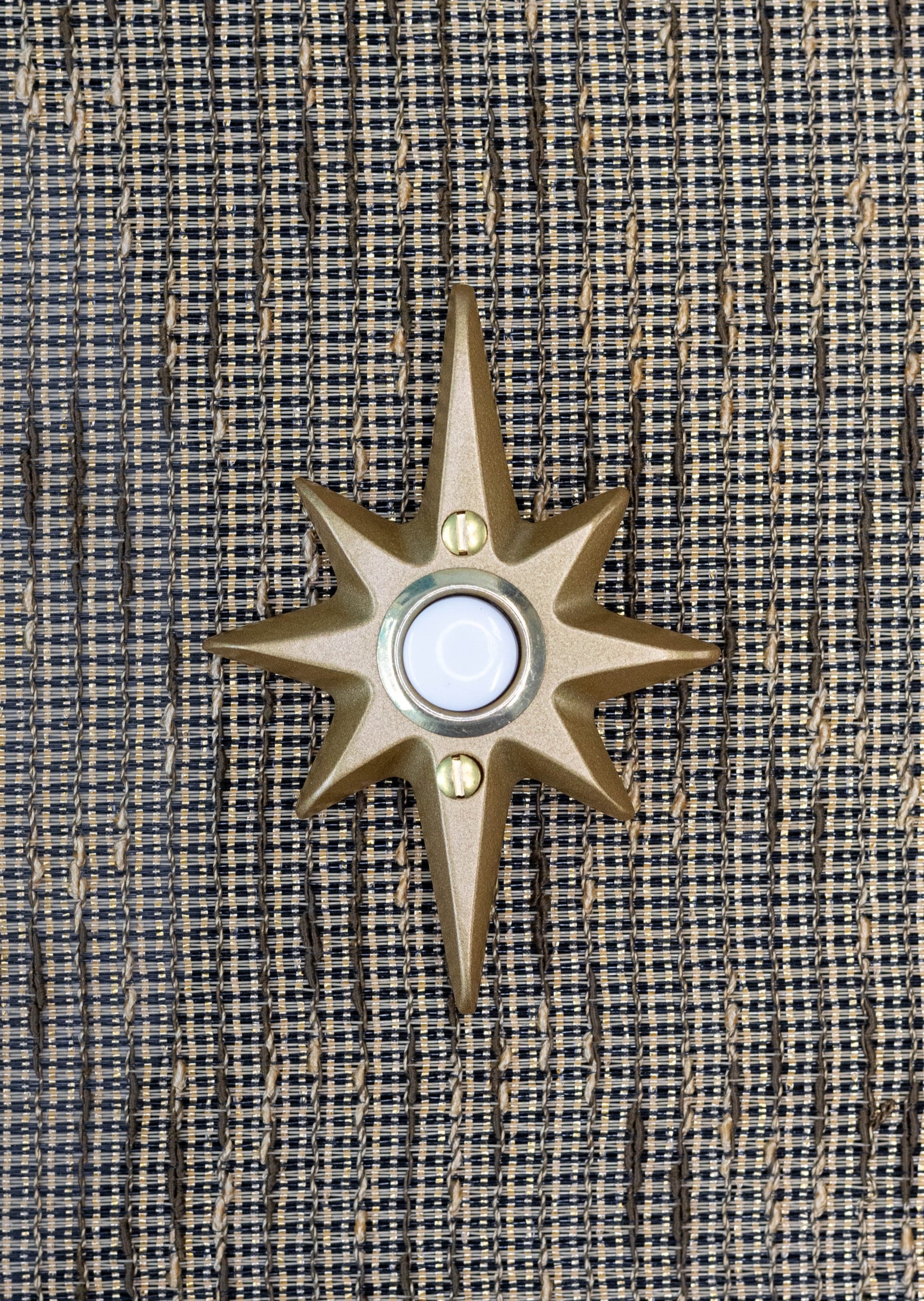 A small gold powder coated cast aluminum starburst with a gold rimmed doorbell in the middle. The doorbell itself is a white plastic. There are two gold screws, one below and one above the doorbell to attach the starburst to the wall. The gold powder coating has a smooth metallic finish that is slightly reflective of light.