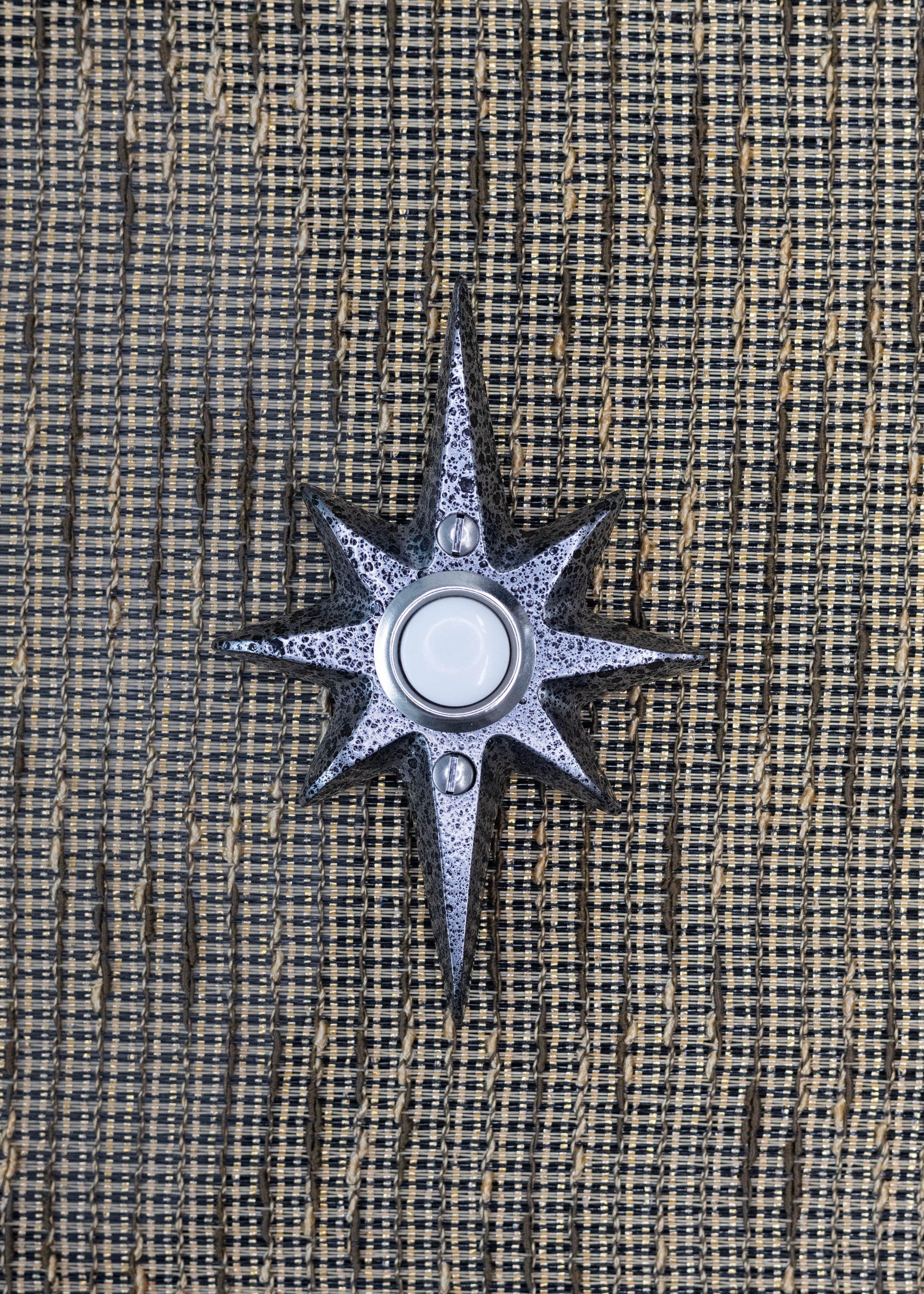 A small black frost powder coated cast aluminum starburst doorbell button. The powder coating is a darker silver with black pitted texture. It has a nice metallic shine to it. The doorbell button is a white plastic with a silver rim. There are two silver screws, one above and below the doorbell.