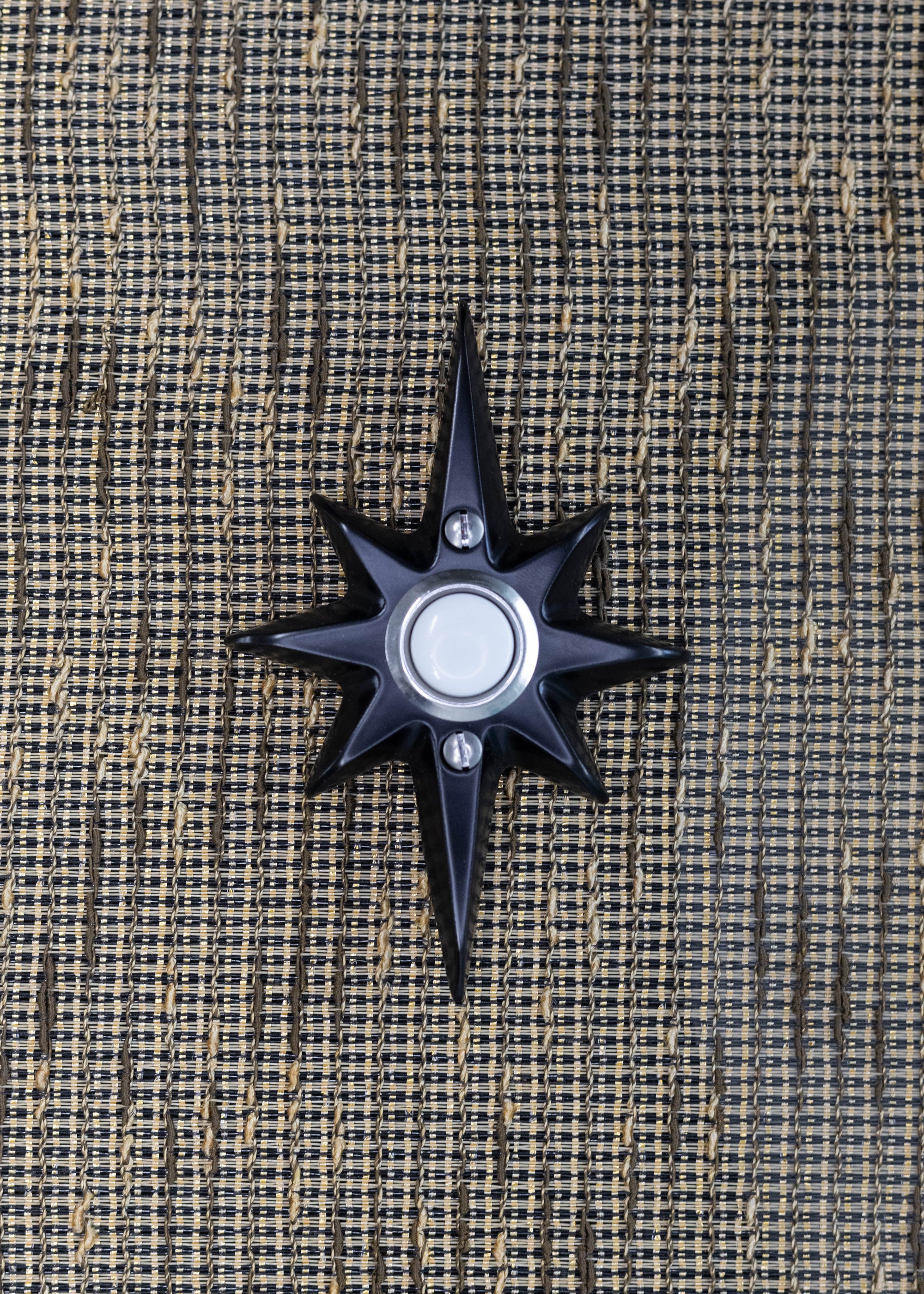 A small black powder coated cast aluminum starburst with a door bell in the center. The door bell is a white plastic with a silver metal rim. There are two silver screws, one above and below the doorbell. The powder coating is very smooth, and reflects a bit of light, but is not glossy.