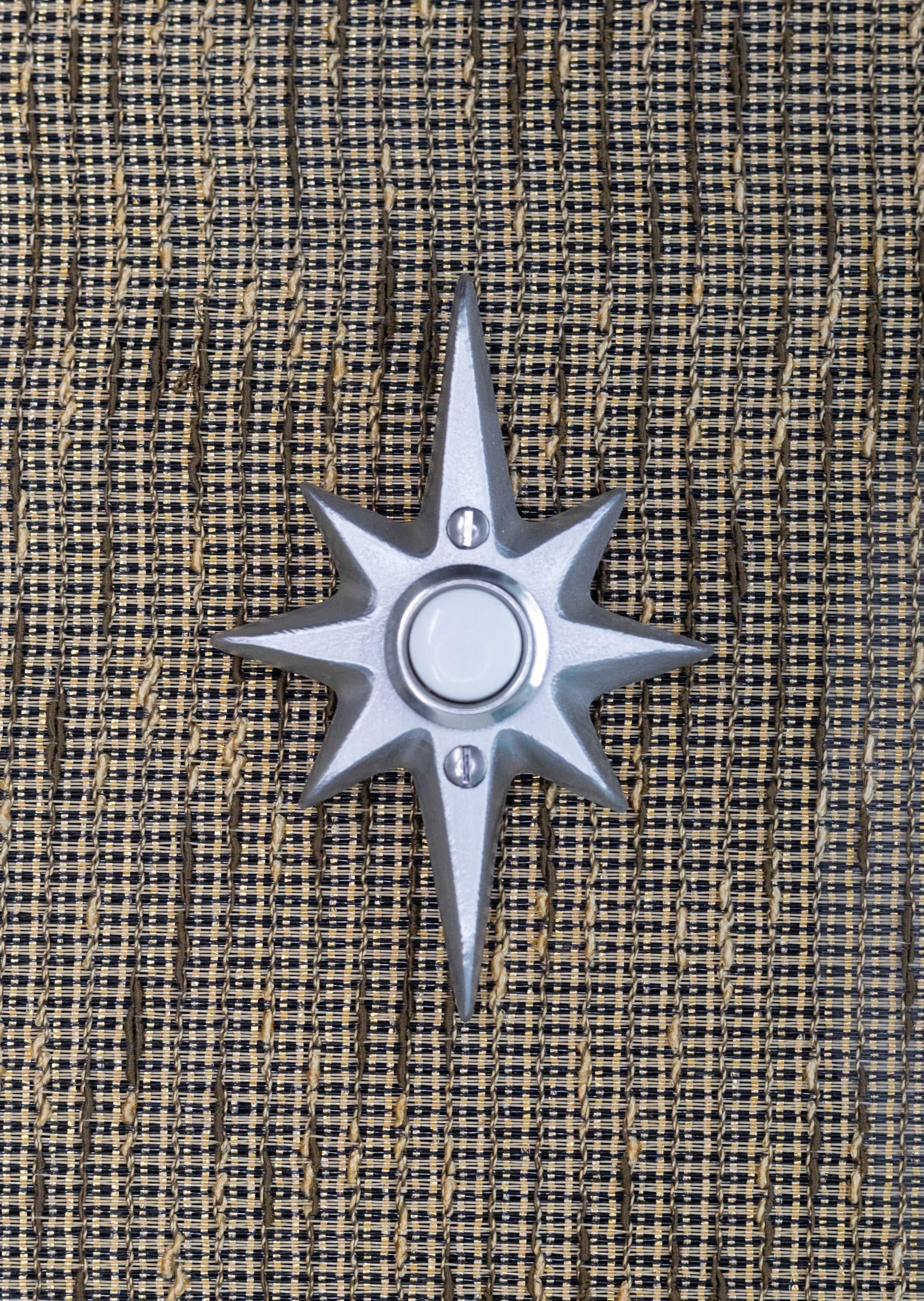 A small natural cast aluminum starburst doorbell. The natural aluminum has a small bit of porous texture to it, but is still very smooth and reflects the light. There are two silver screws, one above and below the doorbell in the center.
