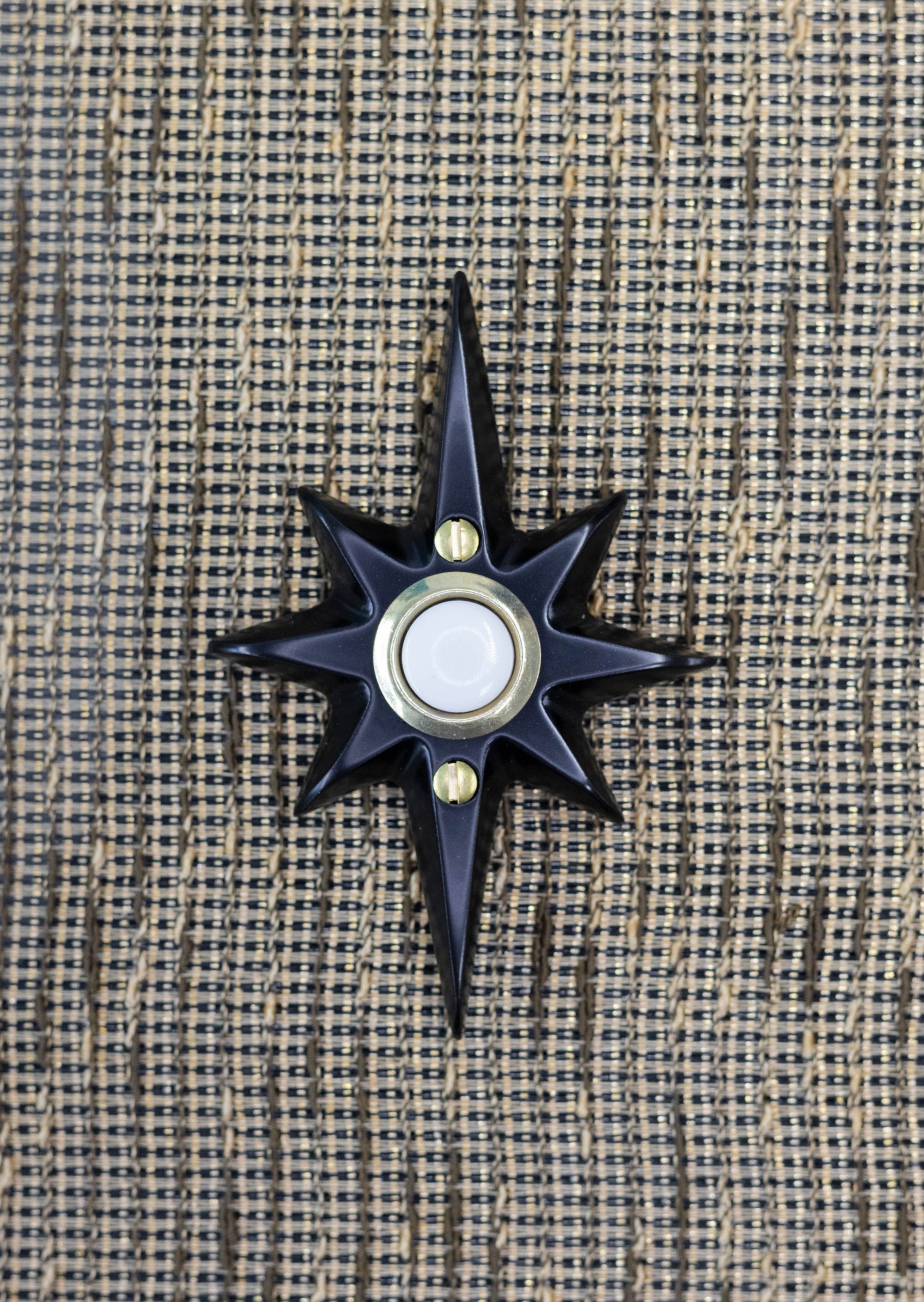 A small black powder coated cast aluminum starburst with a door bell in the center. The door bell is a white plastic with a gold metal rim. There are two gold screws, one above and below the doorbell. The powder coating is very smooth, and reflects a bit of light, but is not glossy.